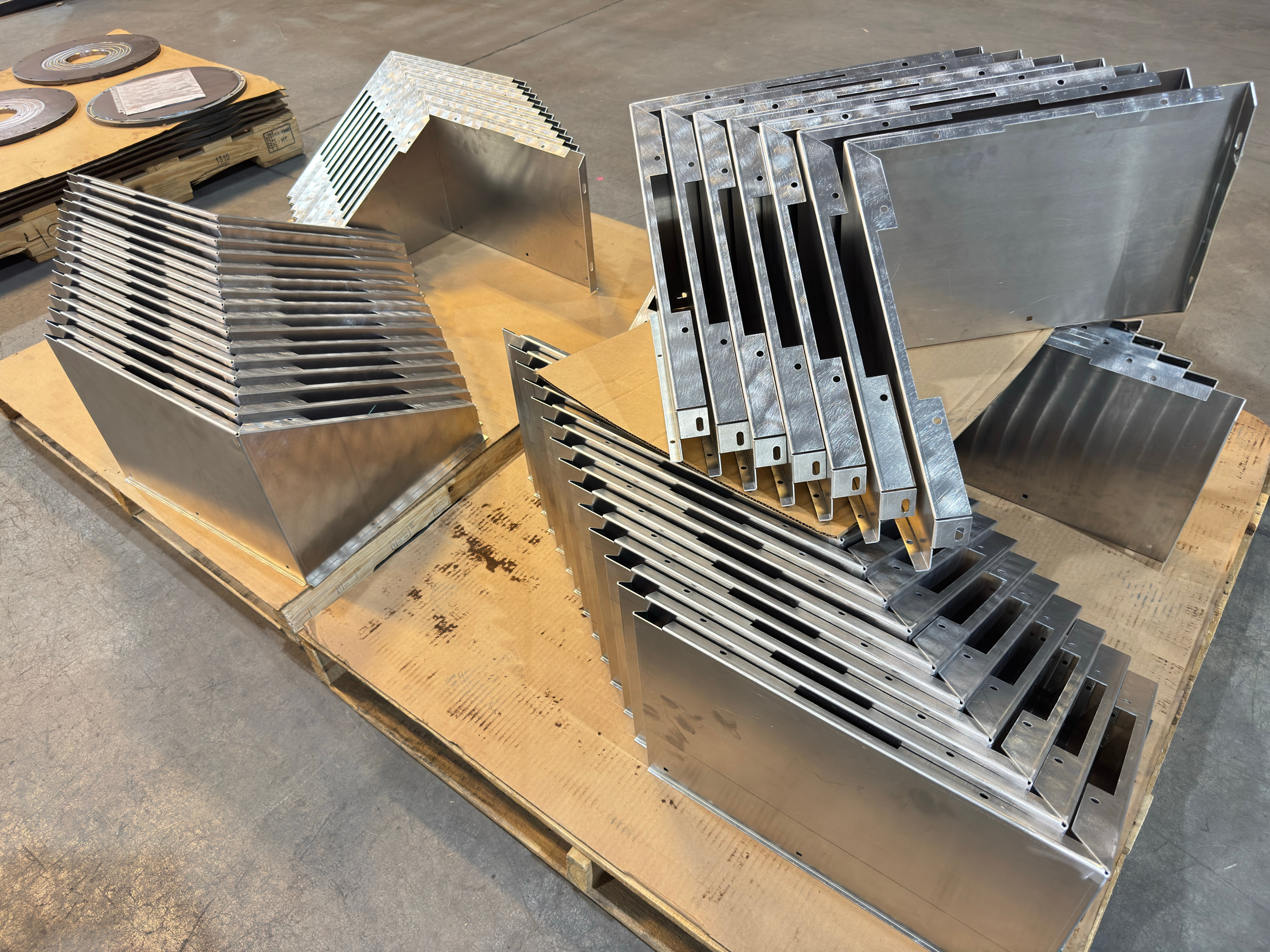Stacks of metal brackets and sheet steel parts arranged on a wooden pallet in a workshop.