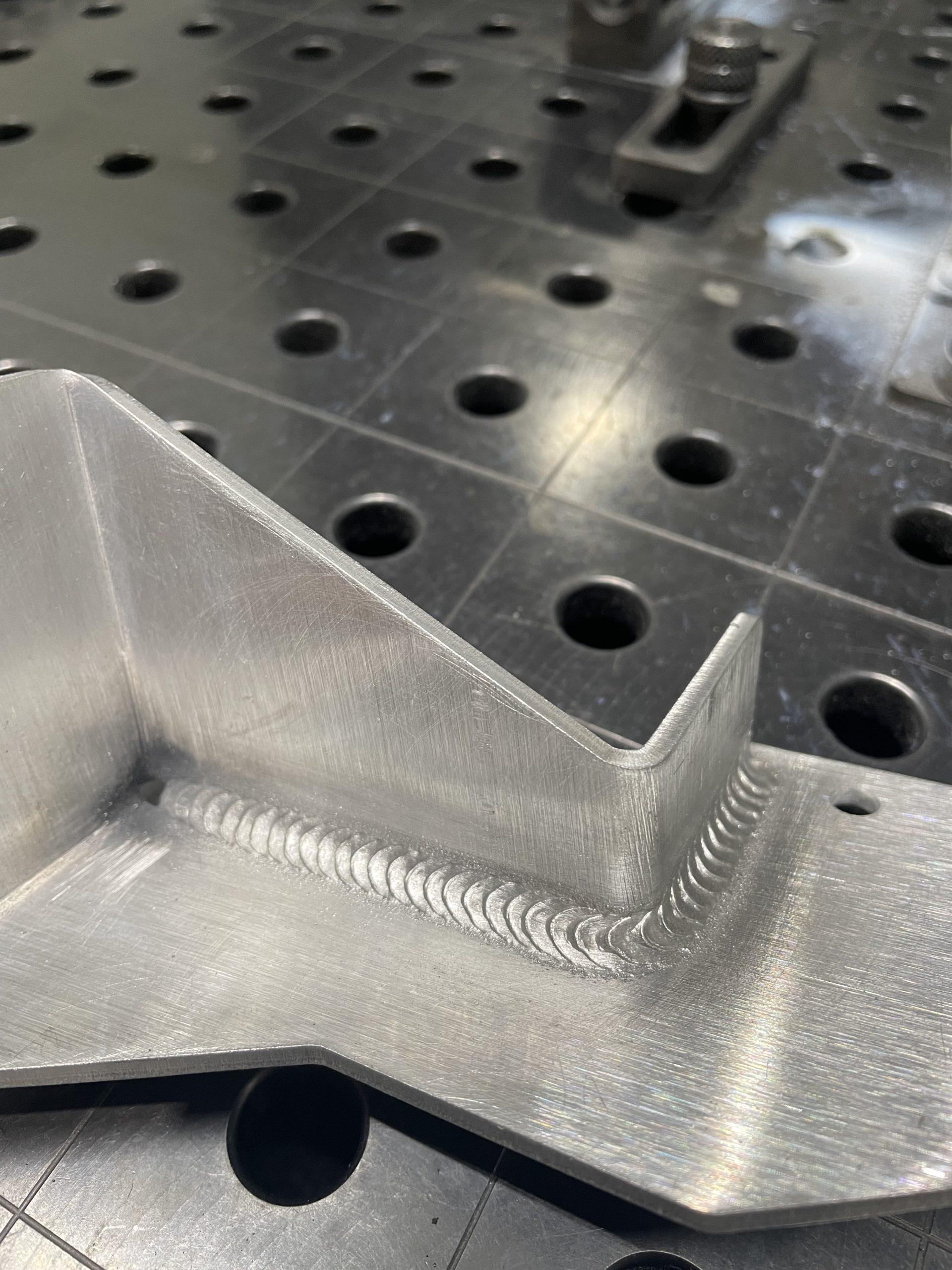 Brushed metal bracket with a welded seam resting on a perforated welding table.