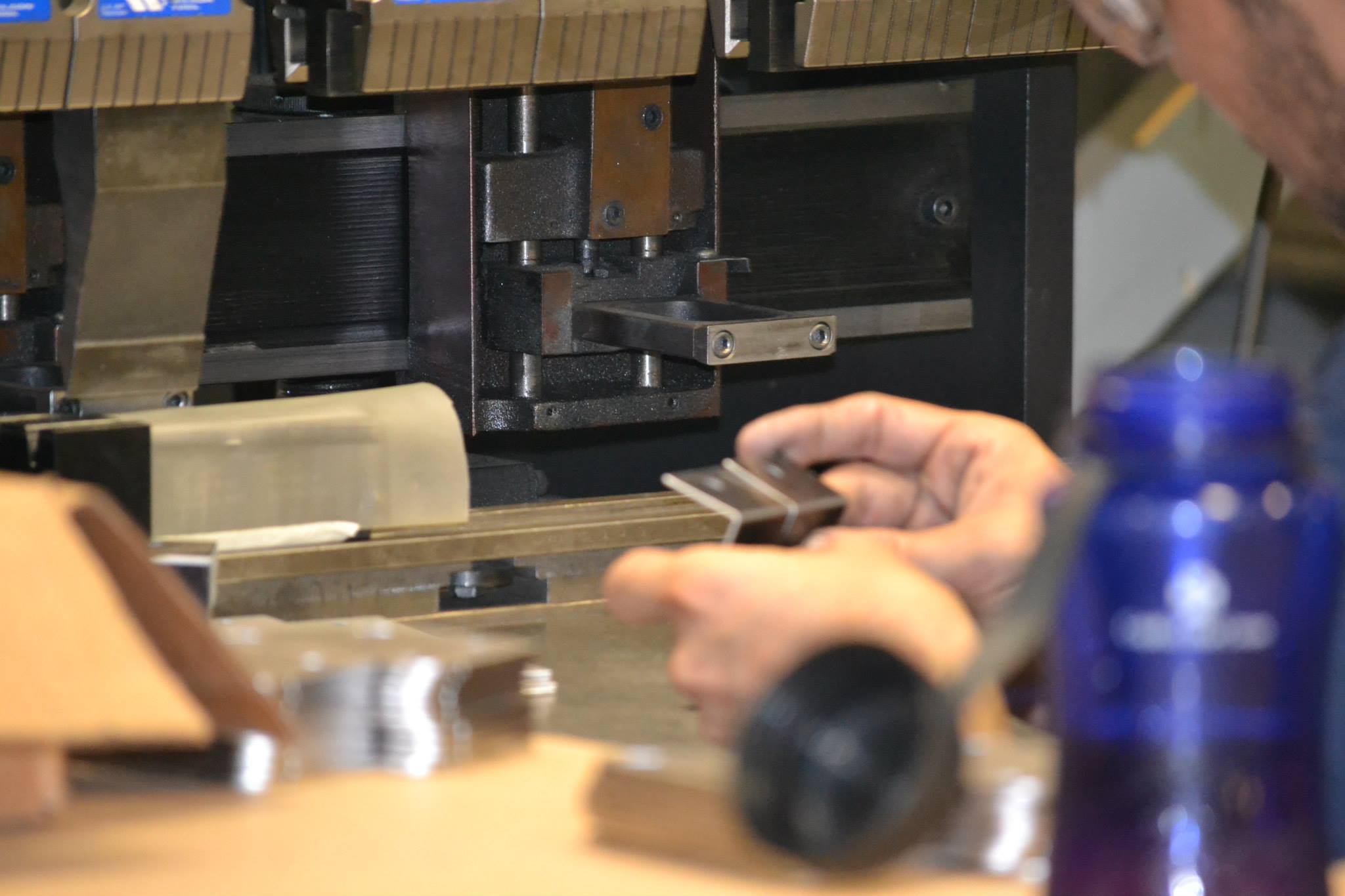 Hands operate a machine tool in an industrial workshop with metal parts and equipment.