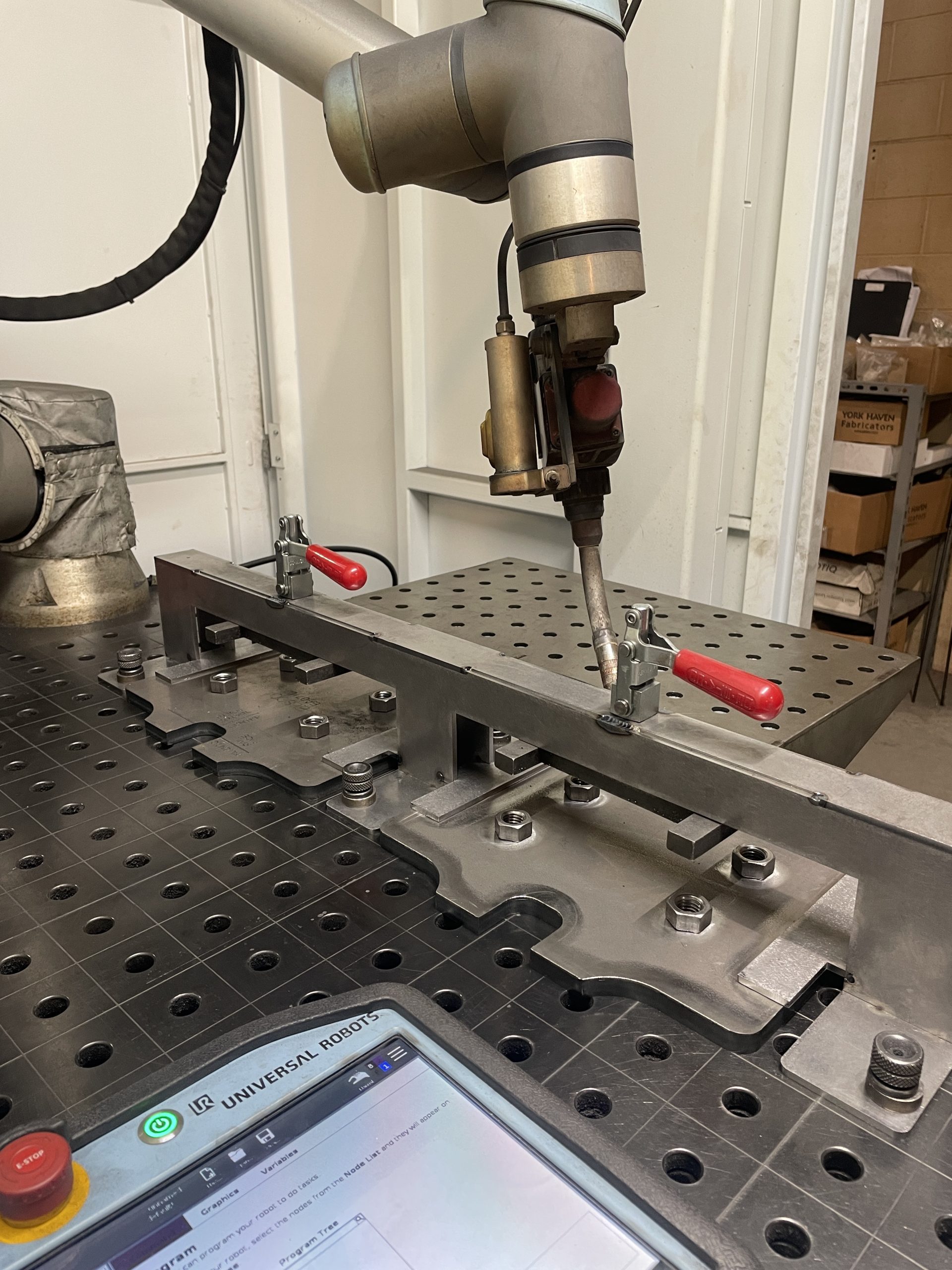 Robotic arm welding a metal assembly on a perforated worktable in an industrial workshop.