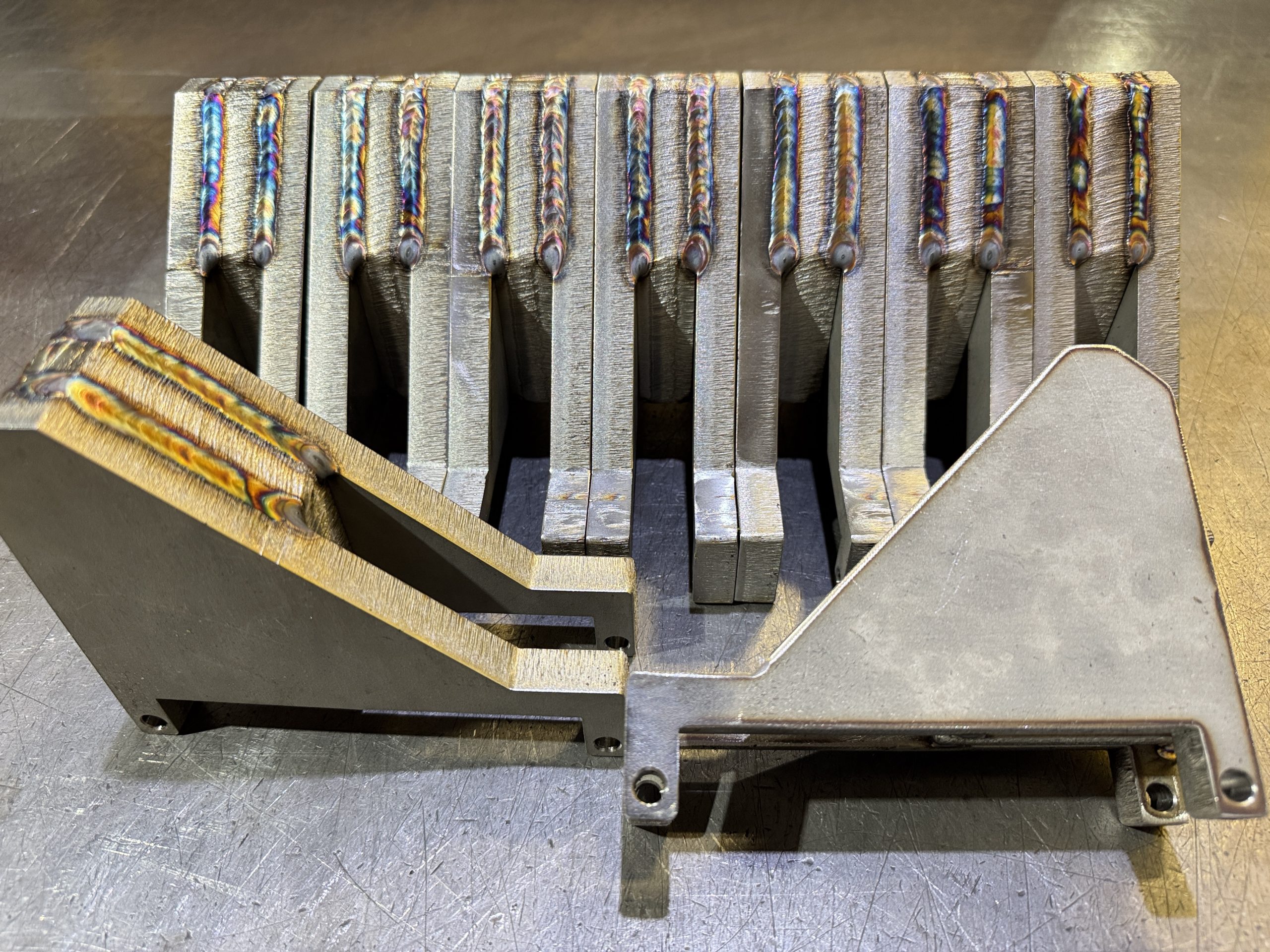 Metal welding jig with multiple slotted clamps arranged on a workbench.